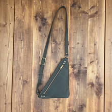 Load image into Gallery viewer, A dark green triangular crossbody bag with a long strap, brass zipper, and brass buckle, laid flat on a rustic wooden plank background.
